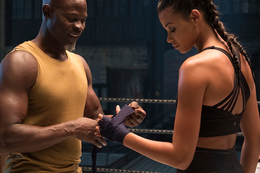 Djimon Hounsou smiles while helping Ella Balinska wrap up left hand with navy boxing hand wrap, both stand in boxing ring.
