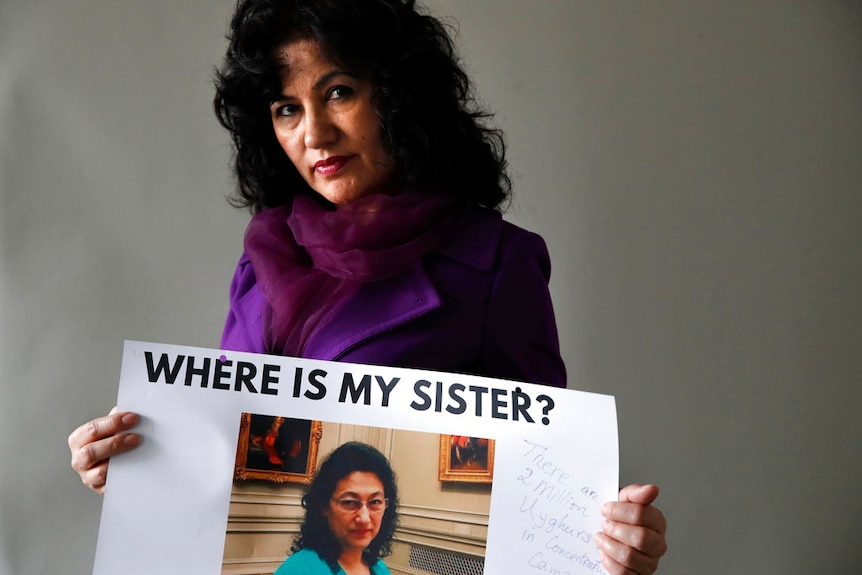 A woman with dark hair wearing a purple coat and scarf holds up a sign with a photo saying 'Where is my sister?'