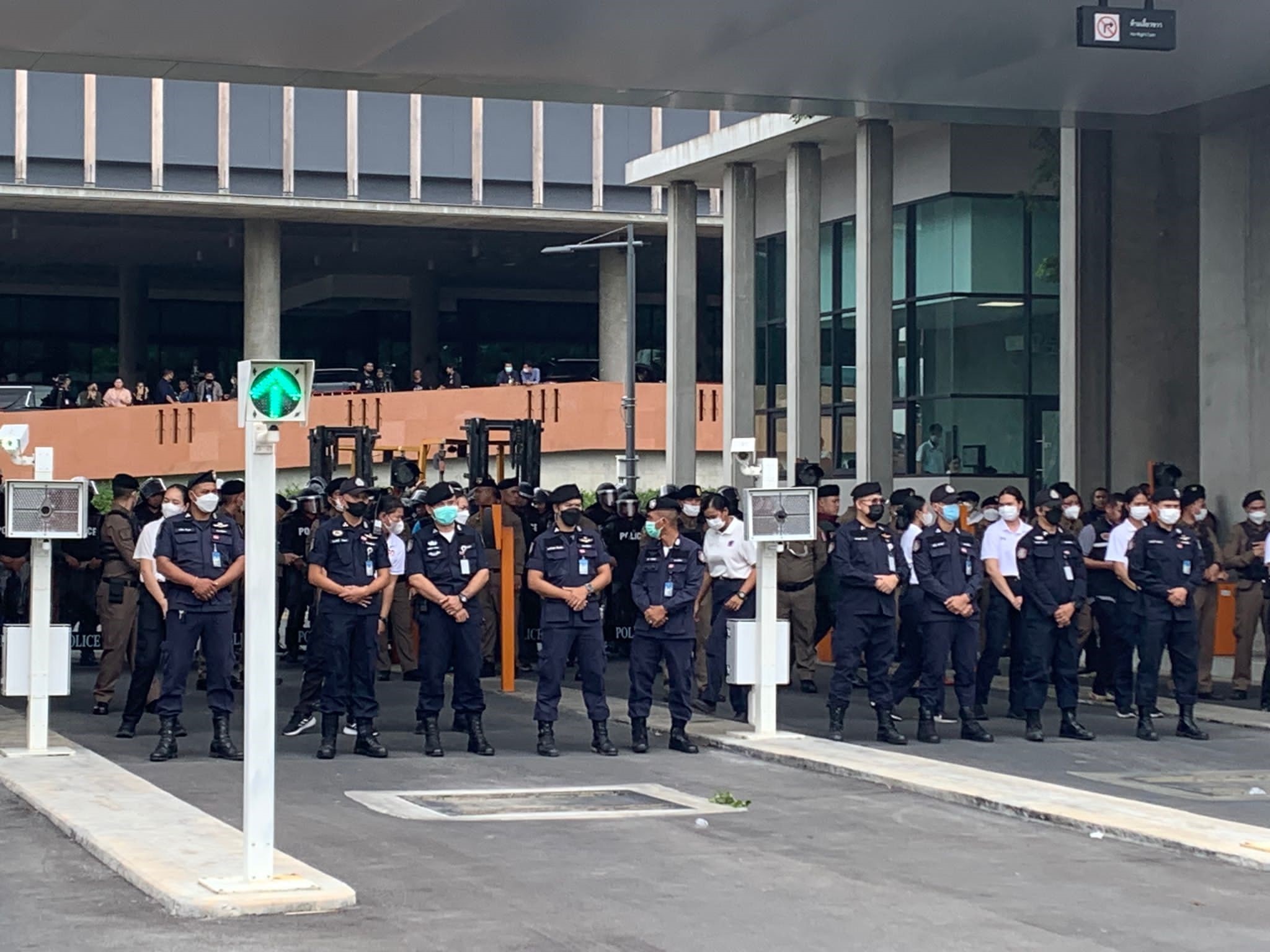 Thai police in dark uniforms stand in lines blocking a road, with large buildings nearby.