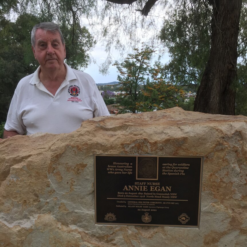An old man in a white polo shirt stands behind a standstone rock with a bronze plaque that reads 'Annie Egan'.