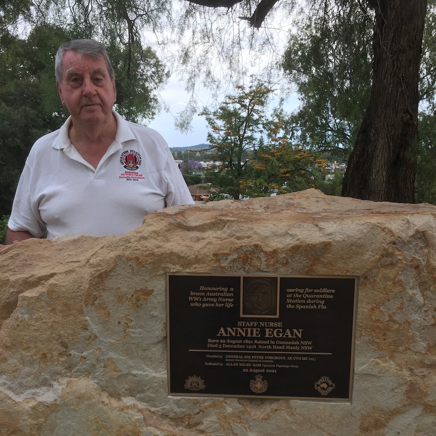 An old man in a white polo shirt stands behind a standstone rock with a bronze plaque that reads 'Annie Egan'.