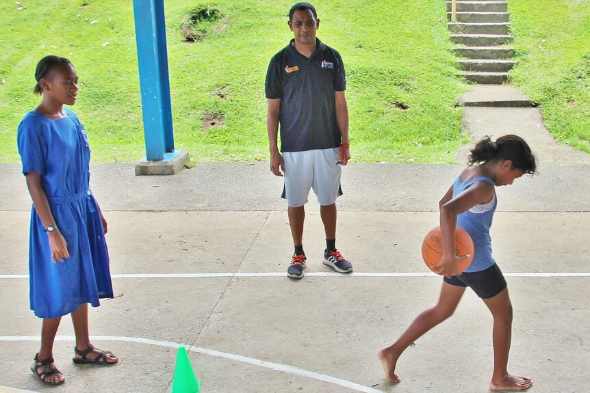 A small girl dribbles a ball up court with a taller girl waiting in line to go next. Kenneth watches on.