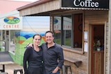 A man and woman stand with their arms around each other outside a small cafe.