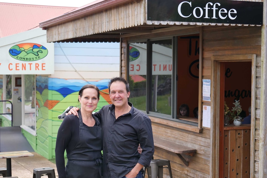 A man and woman stand with their arms around each other outside a small cafe.