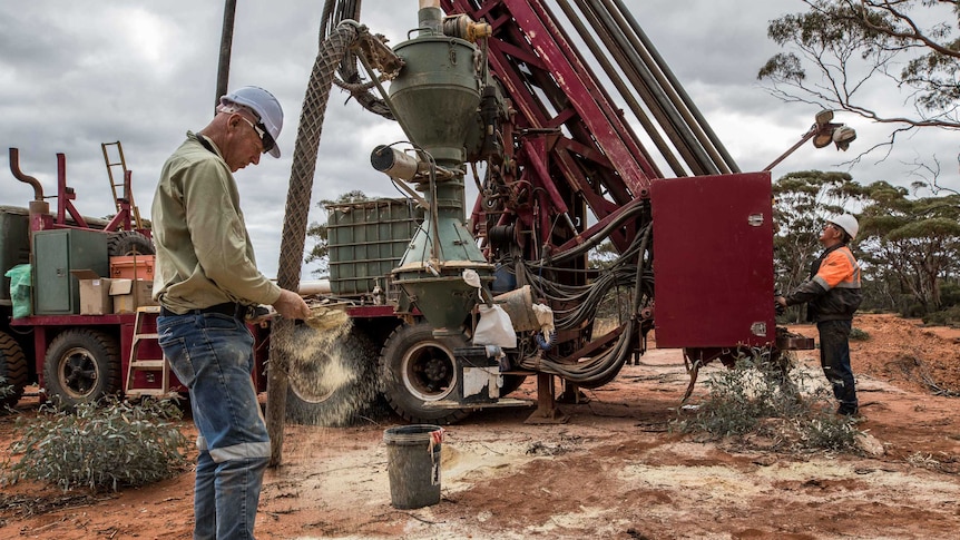 A male geologist sifts through drilling samples next to a drill rig which has a driller at the controls drilling for gold.