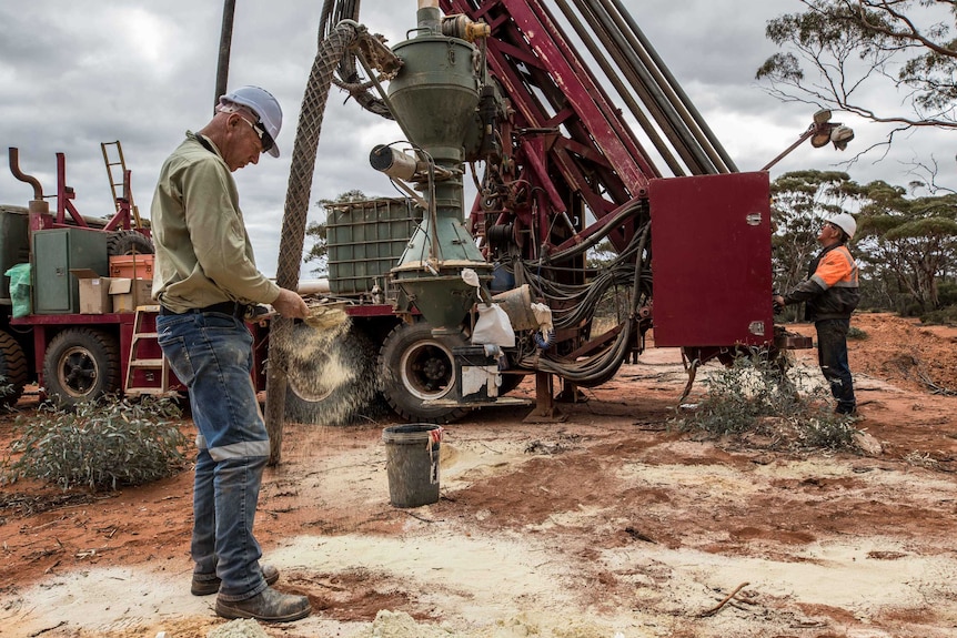 A male geologist sifts through drilling samples next to a drill rig which has a driller at the controls drilling for gold.