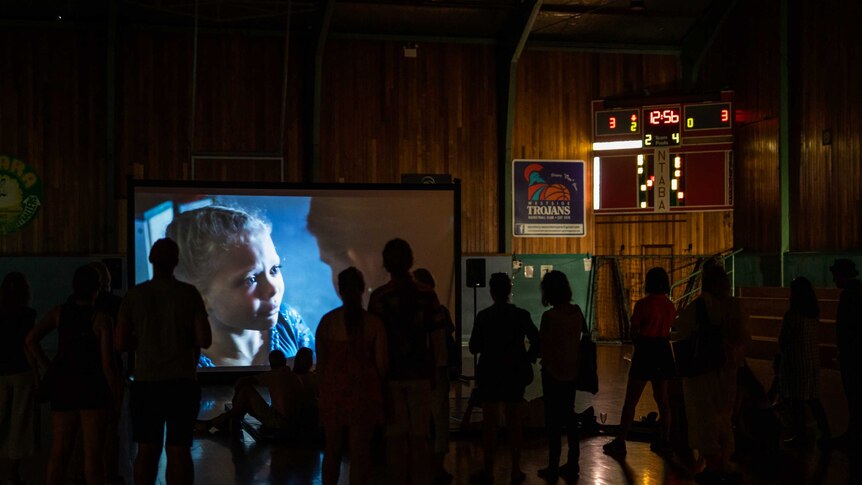 The silhouettes of a crowd of people watching a video art projection in an older wooden basketball court