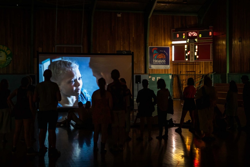 The silhouettes of a crowd of people watching a video art projection in an older wooden basketball court