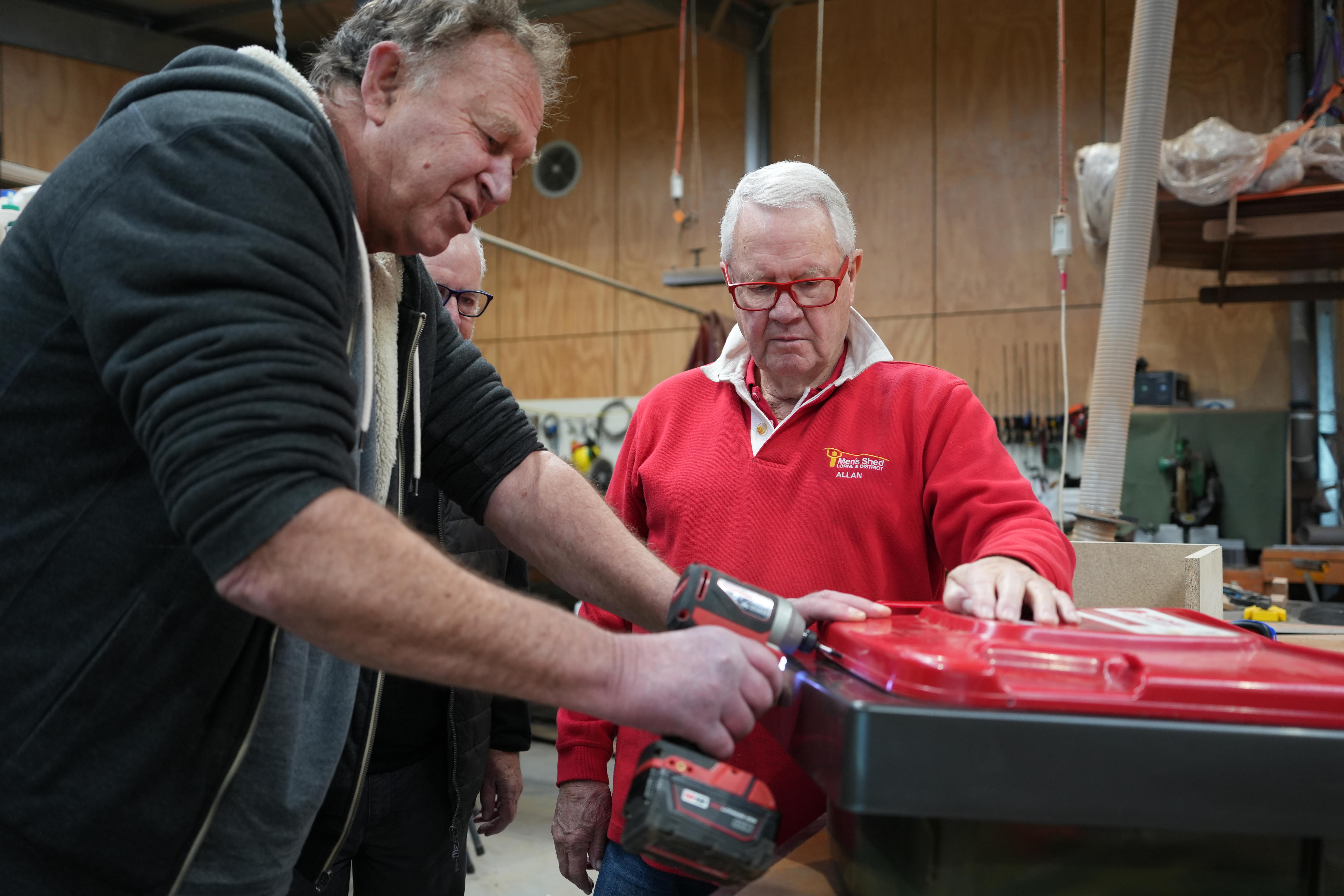 A man with curly hair uses a power tool to secure the lid of a wheelie bin while a man with a red top watches.