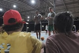 Two men stand in a basketball court with two children sitting listening to them
