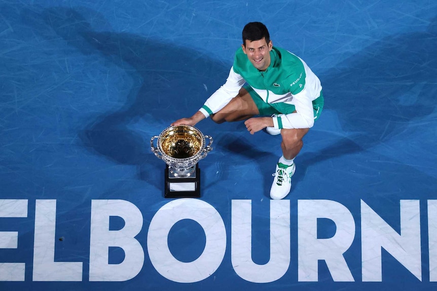 A tennis player looks up as he kneels with the Australian Open trophy and a Melbourne sign.