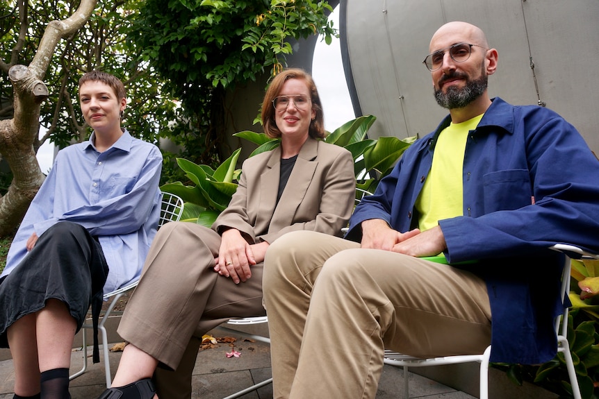 Three people smile while sitting on chairs in front of trees, looking at the camera.