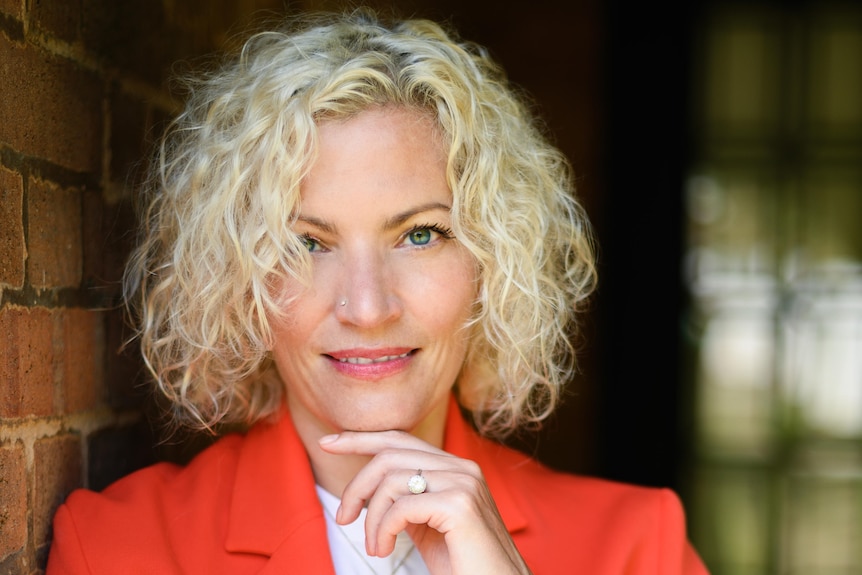 Woman with short, curly blonde hair, wearing red jacket and pants, rests hand under chin, smiling slightly.