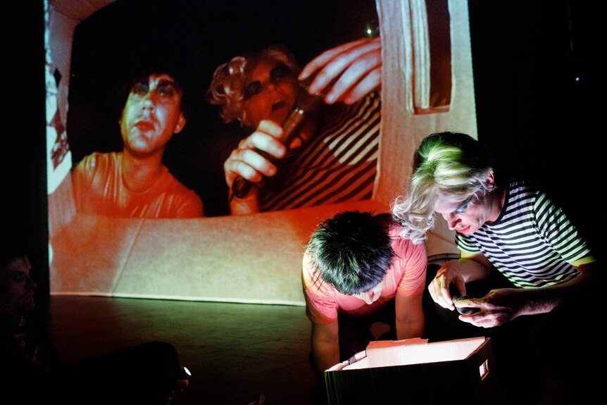 Stage shot, darkened, two men in front of stage stare into cardboard box while their faces are projected onto screen behind.