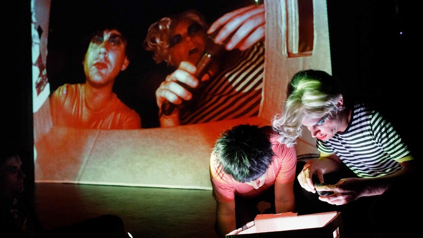 Stage shot, darkened, two men in front of stage stare into cardboard box while their faces are projected onto screen behind.