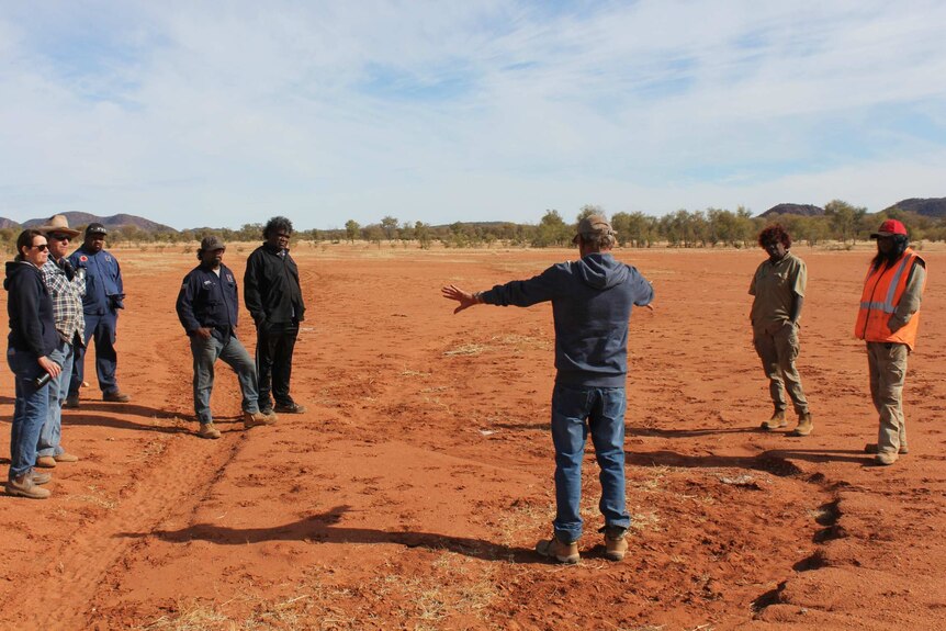 Group of people standing on red dirt listening to a man in the middle whose back is to the camera