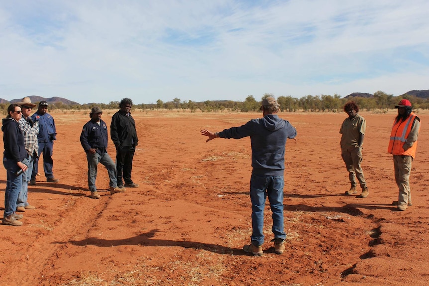 Group of people standing on red dirt listening to a man in the middle whose back is to the camera
