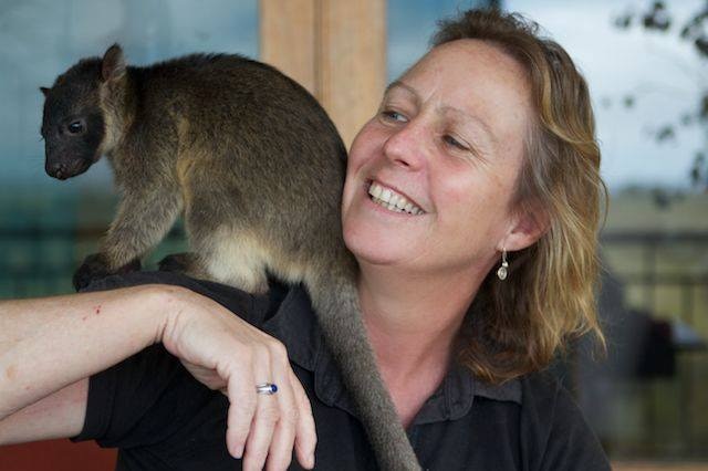 Woman smiles as small furry marsupial with long tail sits on her shoulder.