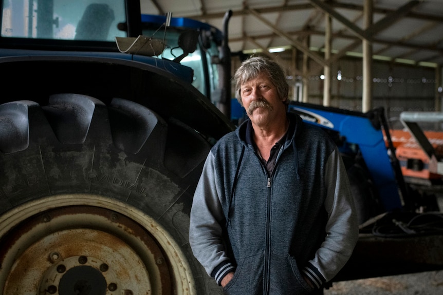 A mustached man in a navy and grey hoody looks into the camera with blue farming equipment behind him.