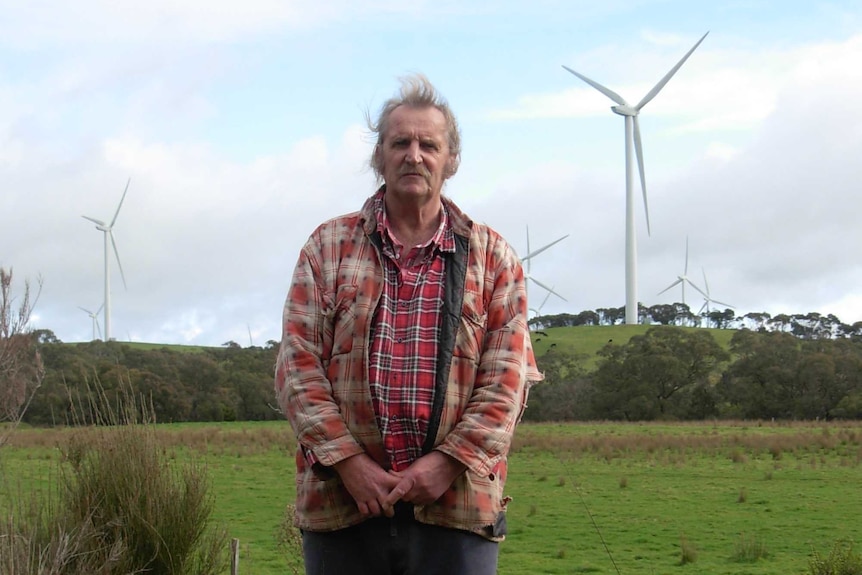 An older man stands in a field with a large wind turbine in the distance behind him.