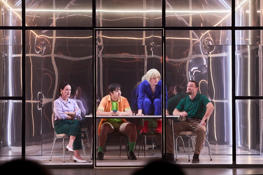 Two women and two men sit around a table inside a large reflective glass house on a stage.