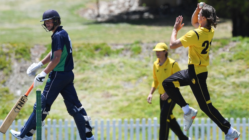 A bowler runs into to bowl wearing yellow and black next to a batter wearing blue.