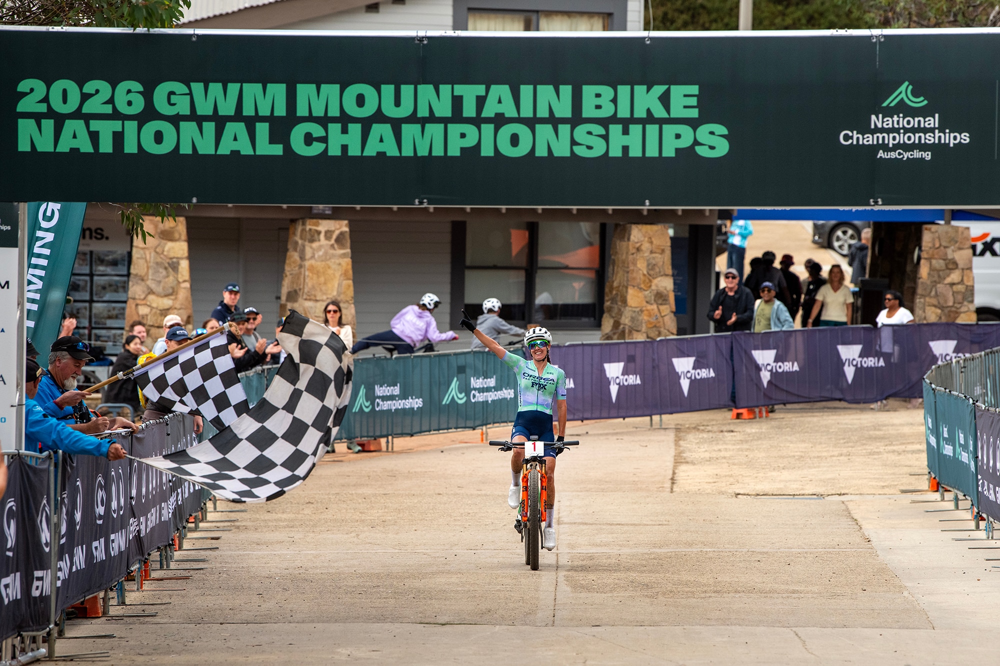 Bec Henderson cruza a linha ao lado de uma bandeira quadriculada e acena com a mão