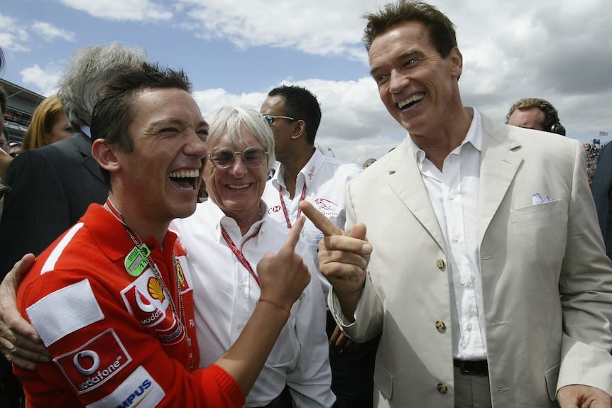 A jockey in a red Formula One top grins and poses alongside movie star Arnold Schwarzenegger at trackside.