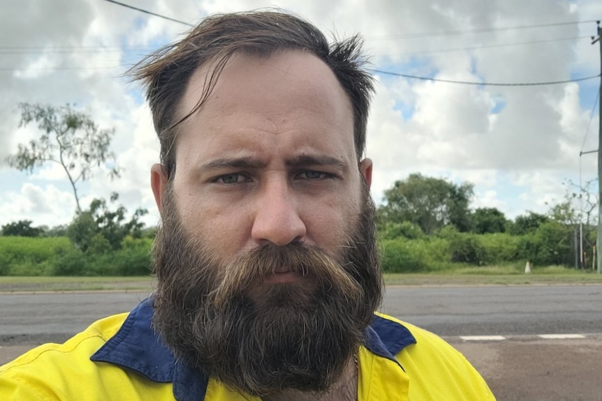Man with dark hair and beard stands in front of road with angered look on his face