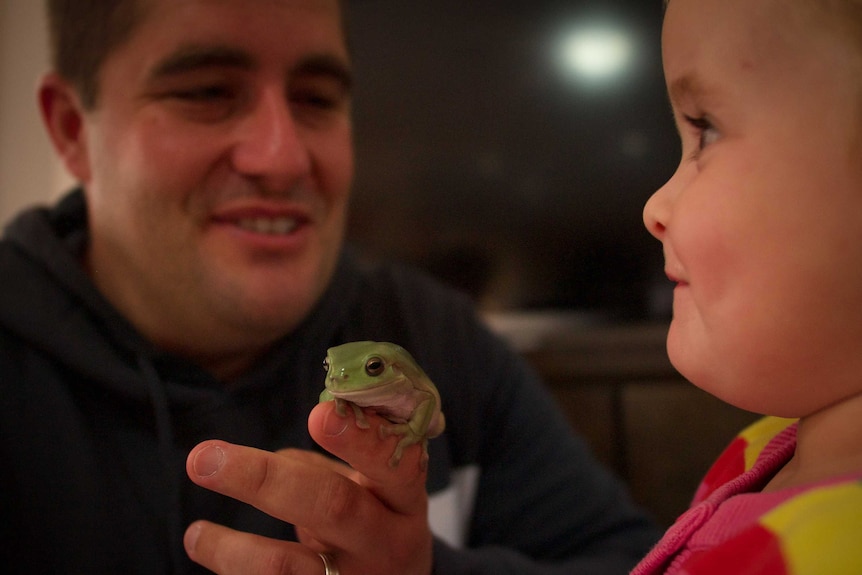 A man holds a green frog up for a child to look at.