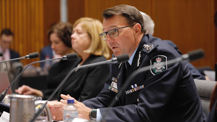 A man in police uniform sits at a desk with a small microphone and nameplate in front of him.