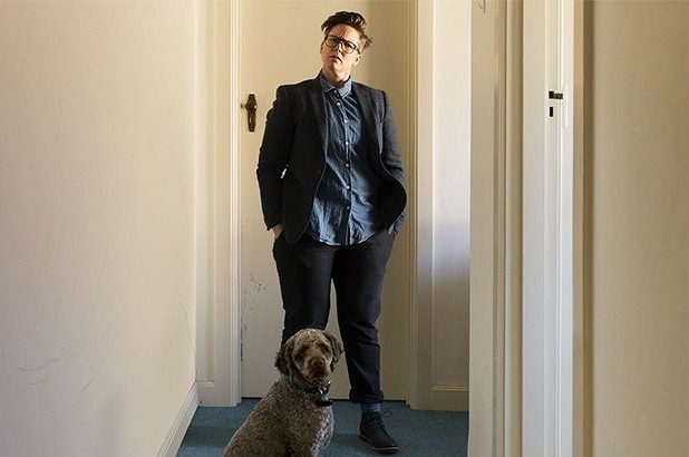 Hannah Gadsby standing by a door in hallway, with her dog Douglas sitting in front of her