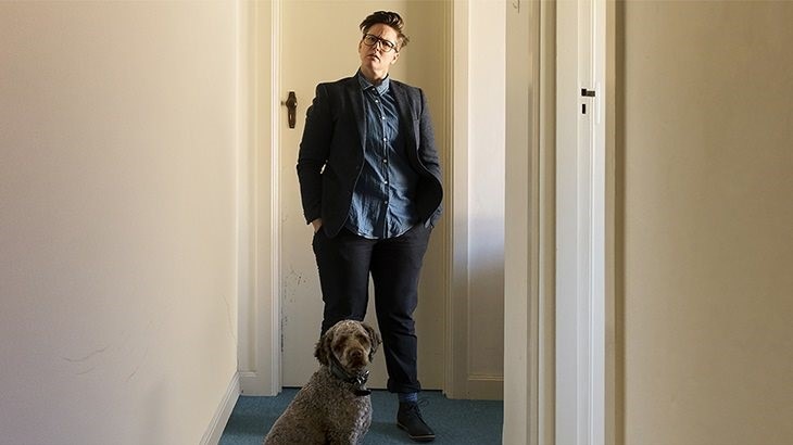 Hannah Gadsby standing by a door in hallway, with her dog Douglas sitting in front of her