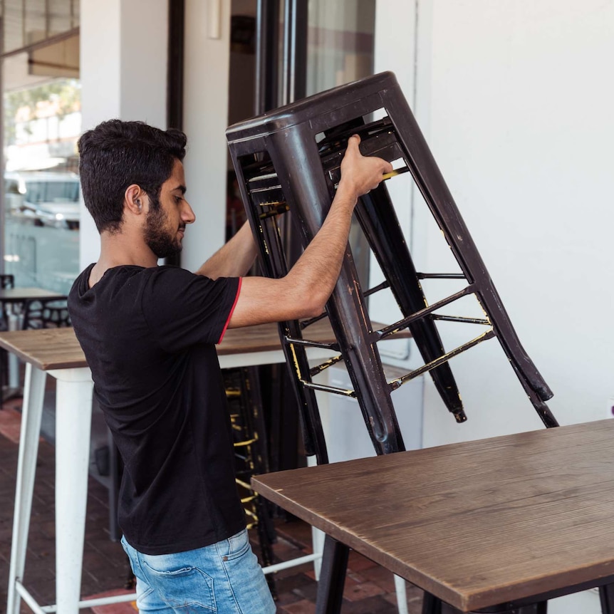 A worker lifts a stack of chairs out the front of the cafe.