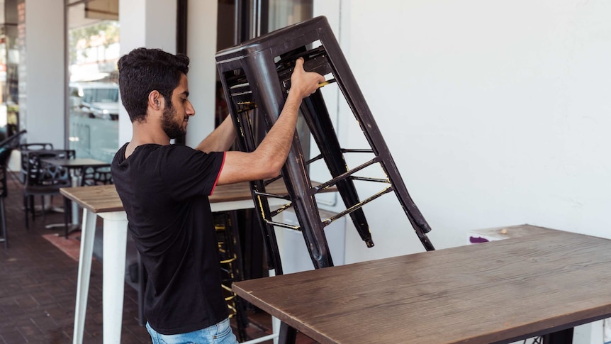A worker lifts a stack of chairs out the front of the cafe.