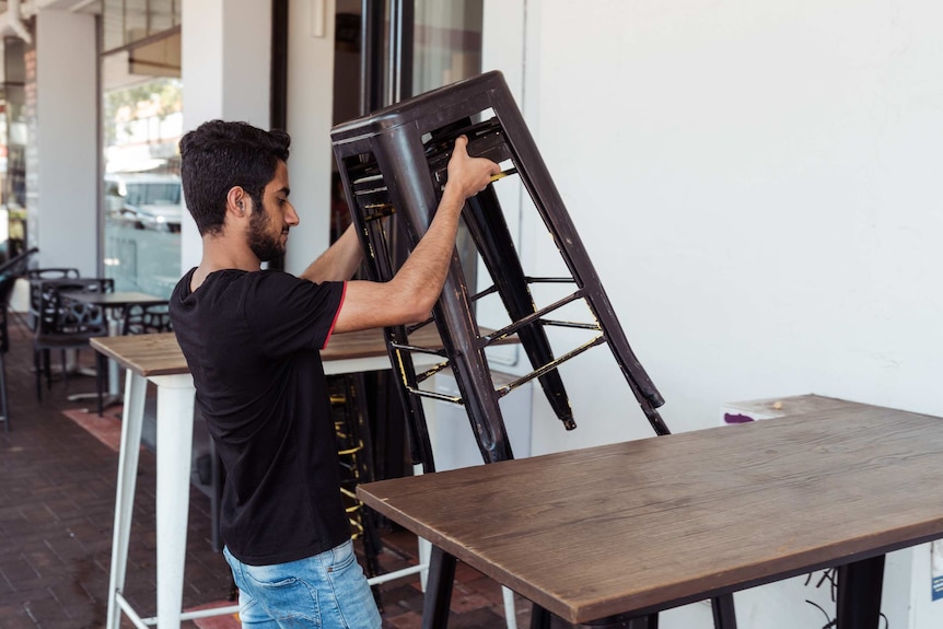 A worker lifts a stack of chairs out the front of the cafe.