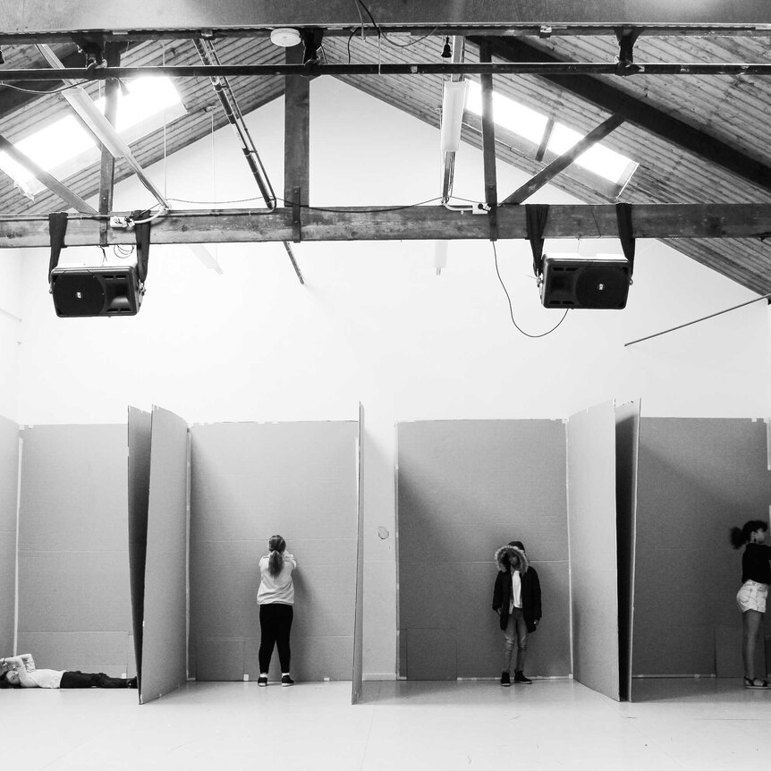 A black and white photo of four children standing in four spaces separated by cardboard in a large room.