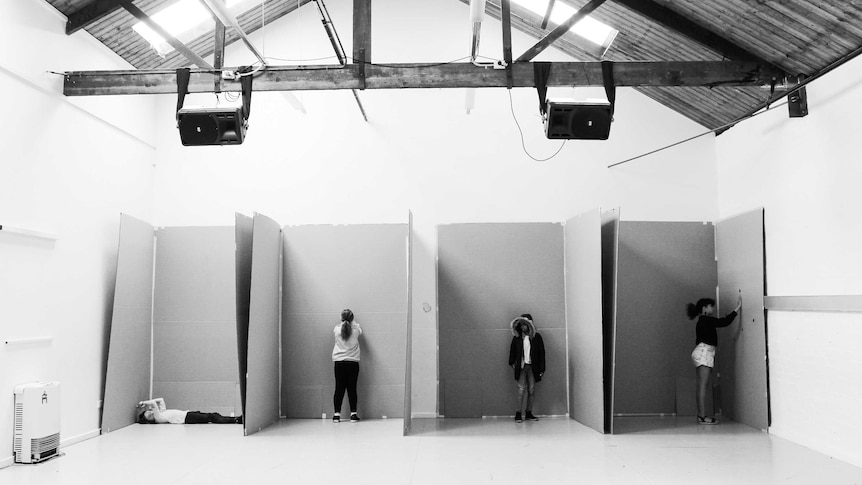 A black and white photo of four children standing in four spaces separated by cardboard in a large room.