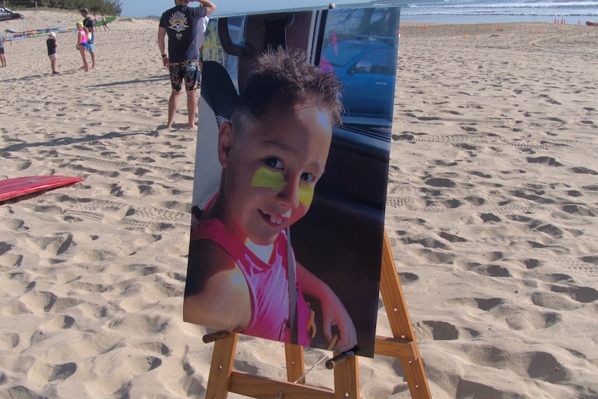 A large photo of a boy in a fluoro nippers vest mounted on an easel on a beach.