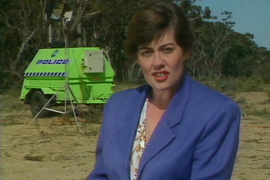 A woman in a purple jacket stands in front of a police truck in the bush.