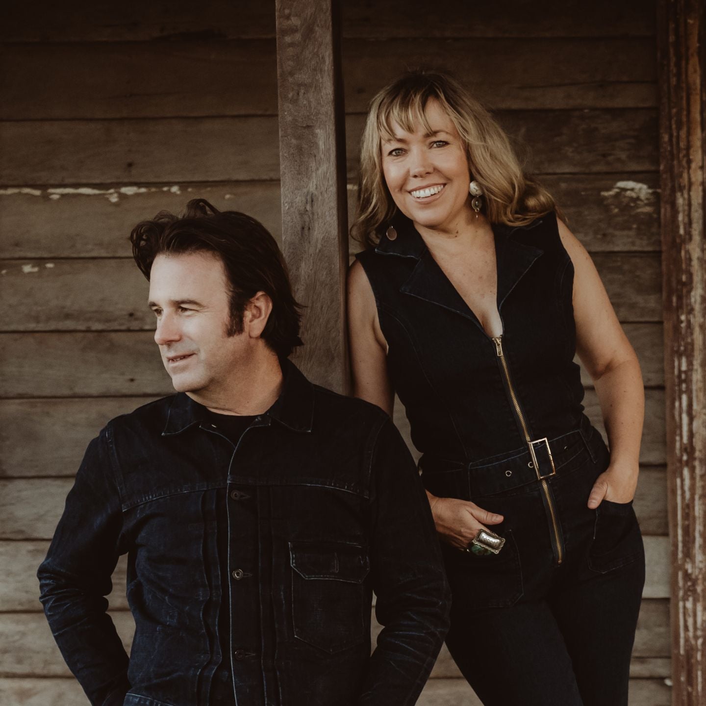 Um homem e uma mulher, ambos vestidos de preto, ficam lado a lado em frente a uma velha casa de madeira.