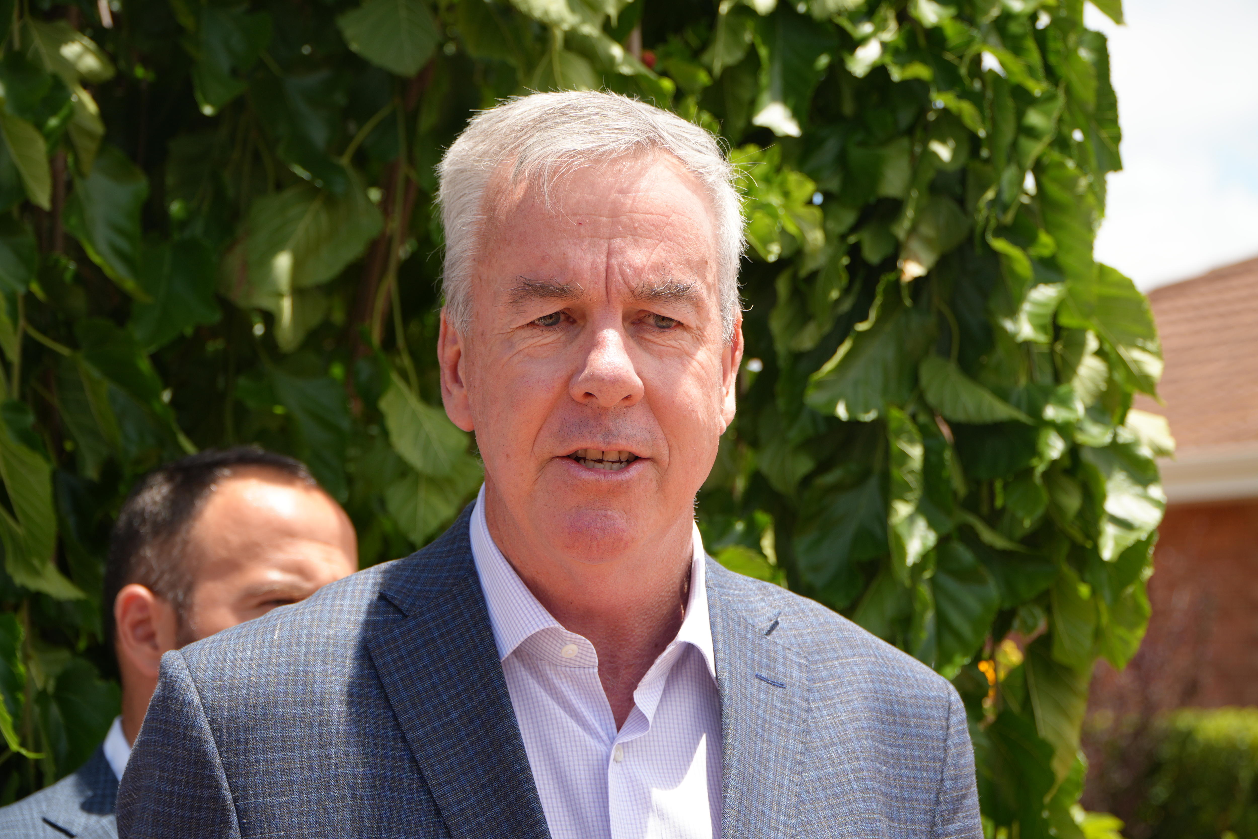 A man with white hair and a grey suit speaks outside at a press conference.