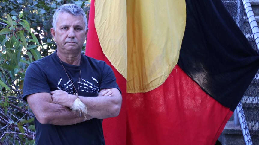 Mr Hayward-Jackson stands with his arms folded in front of an Aboriginal flag. He wears a small tuft of fur on a necklace.