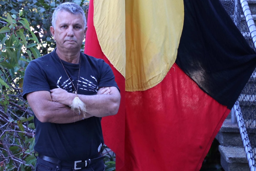 Mr Hayward-Jackson stands with his arms folded in front of an Aboriginal flag. He wears a small tuft of fur on a necklace.