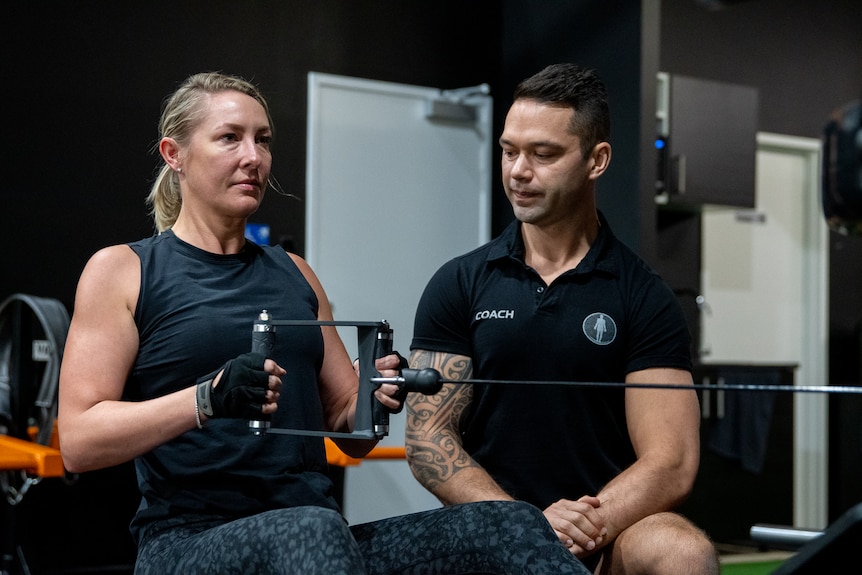 Woman with blonde hair on weight machine under instruction from male trainer