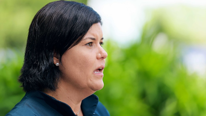 A woman with dark hair stands outside, speaking.