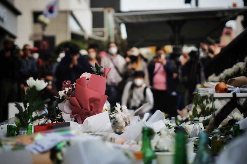 flowers and candles in the foreground, with people in masks gathered in mourning behind