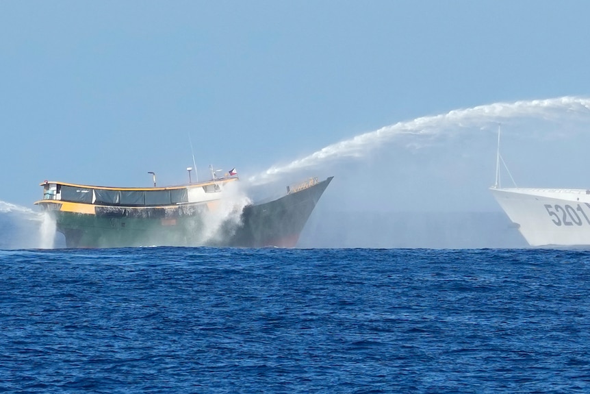 A green boat is hit with a water cannon being shot by a larger white boat in open sea