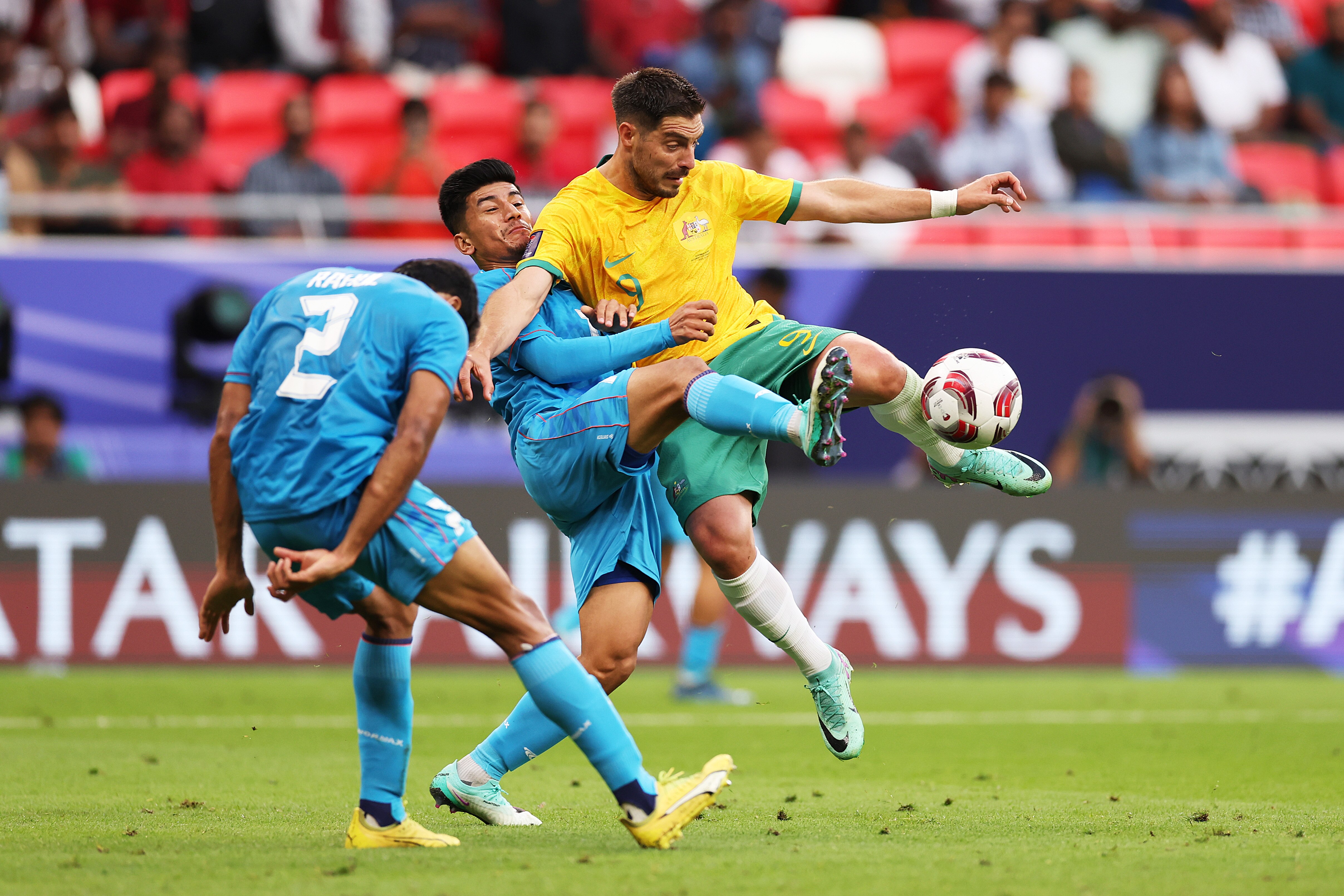 An Indian player crashes into an Australian player as they both raise one leg to kick the soccer ball.