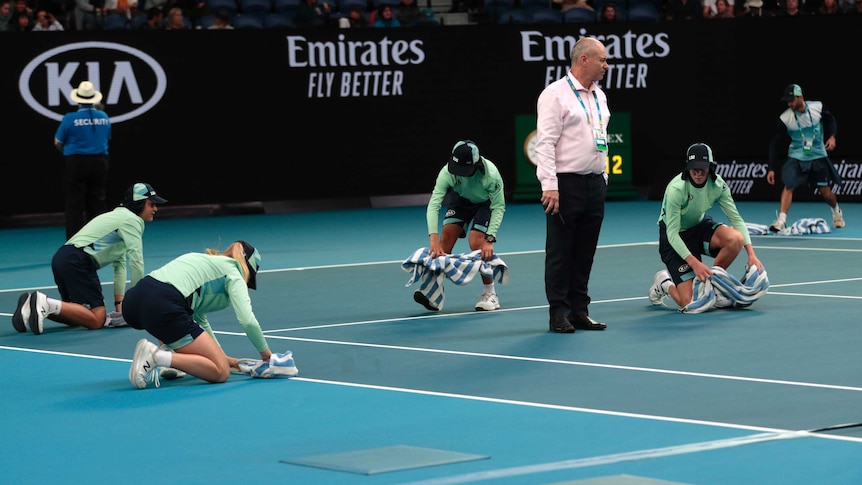 Ball kids can be seen on their knees drying a court with towels whilst a man in a shirt stands in the middle of them